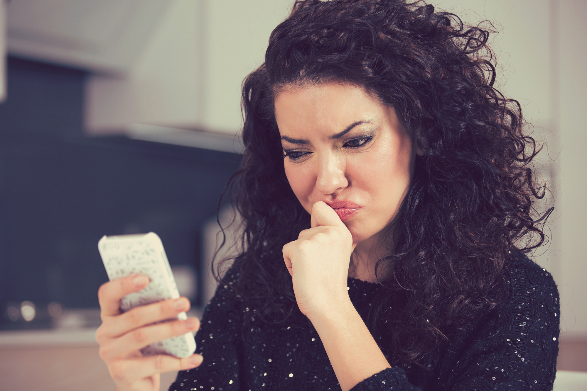 A young woman with long curly dark hair looks skeptically at her mobile phone as she reviews fake dating profiles